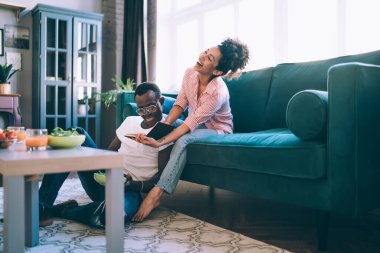 Afro-Amerikalı neşeli bir adam elinde yemekle oturmuş defterine bakıyor ve neşe dolu karısı rahat koltukta dinlenirken gülüyor.