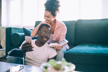 Neşeli, çekici bir eş gülümseyen mutlu kocanın başını telefonla kucaklarken arkada yeşil ev kanepesinde kitapla oturuyor.