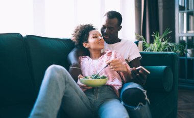 Hoş, çoklu etnik çeşitlilikte bir çift birbirleriyle eğleniyor ve cep telefonu kullanırken kanepede sarılıyorlar ve evde hafif sağlıklı yemekler yapıyorlar.