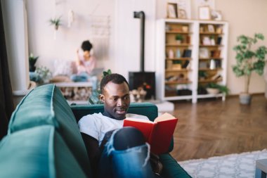 Çok kültürlü, genç bir çift, rahat bir şekilde okuyup yeşil kanepeye ve ferah oturma odasındaki bankta oturmaya odaklandılar. 