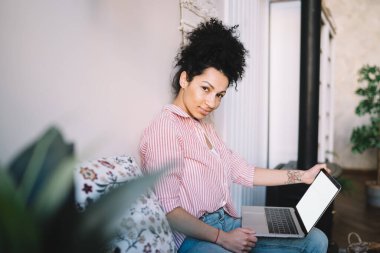 Serene dövmeli, modaya uygun tişörtlü, evinde koltukta dinlenirken dizüstü bilgisayar kullanan ve arka plandaki bulanık kameraya bakan bir kadın.