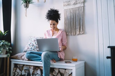 Şen şakrak etek giyen, rahat gömlekli, çapraz bacaklı ve modern odada dizüstü bilgisayar kullanan neşeli bir kadın.