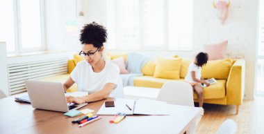 Afro-Amerikalı çocuk kanepede oturup tablette oyunlar oynarken, ebeveynler evde dizüstü bilgisayarda çalışıyor.