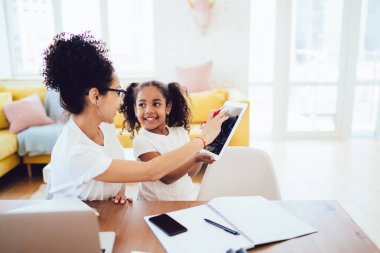 Saçları topuz ve neşeli kıvırcık çocuk tablet masasında parlak odada bulanık arka planda gezen neşeli etnik ebeveynin yan görüntüsü