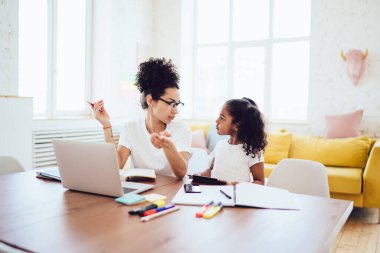 Zeki siyah ebeveyn parlak odada dizüstü bilgisayarın yanında oturuyor ve evde serbest çalışırken tabletini elinde tutan kıvırcık kızıyla konuşuyor.