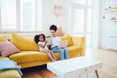 Beyaz gömlekli ve kot pantolonlu Afro-Amerikan anne rahat bir odada kanepede otururken kızlarının tabletine bakıyor.