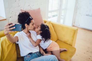 Kıvırcık Afrikalı Amerikalı genç bir bayanın sarı kanepede selfie çekerken tapılası kızını öptüğü yukarıdan görülüyor.
