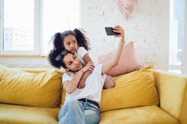 Afrika kökenli genç bir anne cep telefonuyla selfie çekiyor. Şirin, ciddi bir kızı var. Annesinin arkasında, rahat sarı kanepe yastığında oturuyor ve sarılıyor.