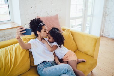 Yukarıdan siyah, genç ve gözleri kapalı bir anneden. Küçük tatlı kızı alnından öpüyor ve evdeki kanepede selfie çekiyor.
