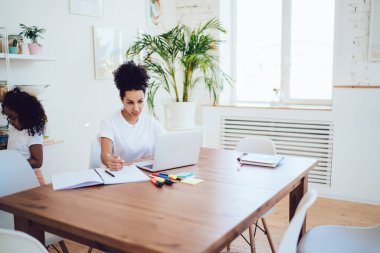 Hafif mutfakta dizüstü bilgisayarda çalışan ciddi etnik bir kadın ve küçük kızı evde geziniyor. 