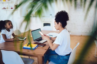 Modern etnik anne sörf telefonunun yan görüntüsü uzaktan bilgisayarla çalışırken evde tablet kullanan küçük bir kızla masada oturmak. 