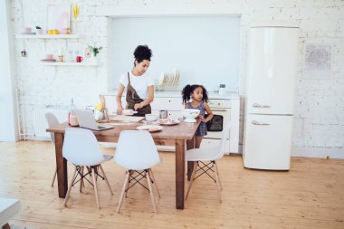 Anne ve kızı, yemek pişirirken yemek masasının önünde dizüstü bilgisayar ve malzemelerle duruyorlar.