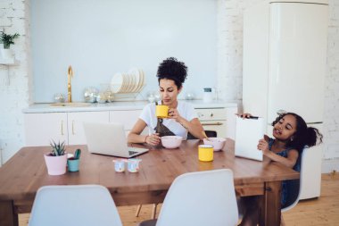 Etnik küçük kız annesiyle tablet izliyor dizüstü bilgisayar izliyor evdeki modern mutfaktaki masada bir bardak içki içiyor.
