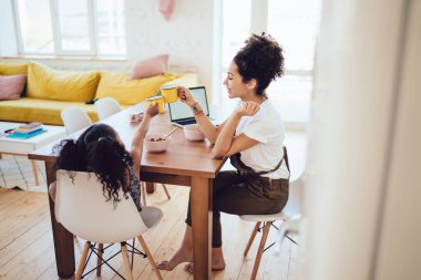 Genç etnik kıvırcık kadın masada oturuyor ve küçük kızı sarı fincanlarla tıkırdıyor evde kahvaltı yapıyor.  