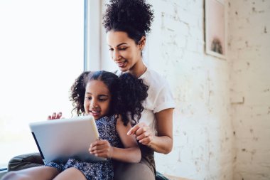 Beyaz tişörtlü Afro-Amerikan esmer anne evde giydiği güzel kıvırcık kızıyla oturuyor ve pencerenin yanında tablet kullanıyor.