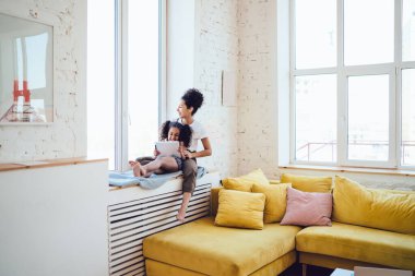 Beyaz tişörtlü kıvırcık saçlı esmer anne, evde mutlu bir kız çocuğuyla pencerenin kenarında oturmuş gülüyor.