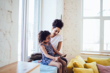 Beyaz tişörtlü kıvırcık Afrikalı Amerikalı annenin, esmer kızıyla pencere eşiğinde oturup, evde birlikte tablet kullanışının yan görüntüsü.
