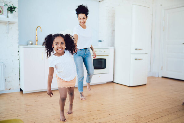 Радостная афроамериканская девушка с вьющимися волосами ходит по гостиной современной квартиры и ведущая мать, глядя в камеру