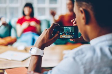 Afrika kökenli Amerikalı bir erkeğin iş arkadaşlarının akıllı telefonuyla fotoğraf çekerken modern ofisin aydınlık kısmında oturması. 