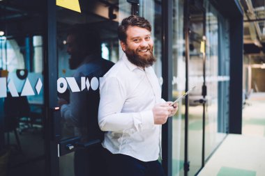 Modern ofis binasındaki cam kapıya yaslanırken kameraya bakıp tablet üzerinde çalışan mutlu erkek ofis çalışanı.