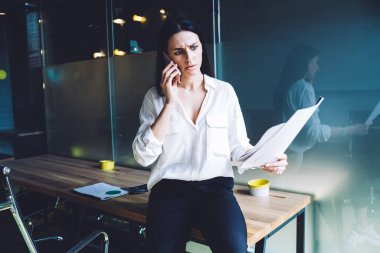 Kadın çalışanın elinde kağıtlar, modern ofis odasındaki masada otururken akıllı telefondan iş ortağıyla konuşması hayret verici.