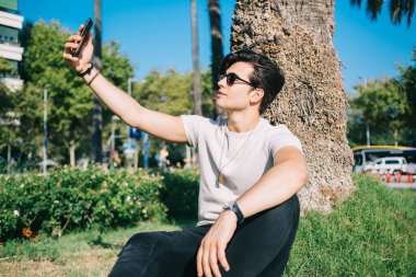 Modayı takip eden beyaz tenli yakışıklı hippi elinde akıllı telefon, parkta selfie çekimi için poz veriyor. Sosyal ağlarda paylaşmak için video çeken genç bir erkek blogcu.