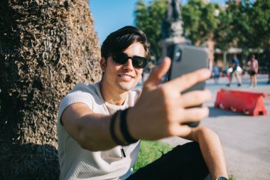 Neşeli 20 'li yaşlarda yakışıklı bir erkek hafta sonları akıllı telefon kamerasıyla selfie çekerek güneşli bir günün tadını çıkarıyor ve cep telefonunda video çekmek için uygulama kullanıyor.