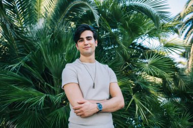 Yeşil palmiye yaprakları üzerinde dikilen günlük tarz giyinen neşeli beyaz erkek portresi. Mutlu 20 'li yaşlarda, haftasonları boş zamanlarında kameraya bakan hippi gezgin.