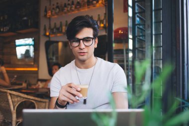 Ciddi yakışıklı adam dijital göçebe kahve içiyor ve dizüstü bilgisayarda video seyrediyor. Kafenin içinde uzaktan çalışıyor. Düşünceli hippi adam internet kursuna Webinar bardakla çalışıyor.