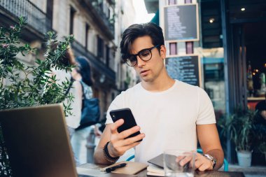 Kafkasyalı ciddi bir beyaz erkek, kafeterya terasında uzaktan çalışırken cep telefonuyla gelir mesajlarını kontrol ediyor. Düşünceli, zengin bir adam blogcusu, internet sitesinde medyayı paylaşmak için modern teknolojileri kullanıyor.