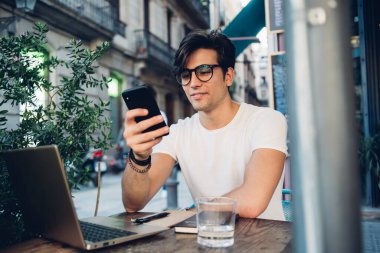 Gülümseyen yakışıklı erkek nüfuzlu kişi, akıllı telefon 4G internet bağlantısıyla sosyal ağlarda içeriği paylaşıyor. Kafe terasında dizüstü bilgisayar ve cep telefonu kullanarak çalışan gülümseyen hippi adam serbest çalışıyor.