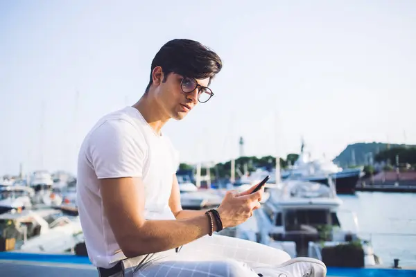 Handsome caucasian hipster guy in spectacles sitting in port using mobile phone for blogging during journey, pensive 20s male tourist chatting via roaming connection sending mail on smartphone