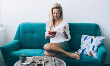 Kaygısız hippi kız evde rahat bir kanepede oturup diyet yemeğinin tadını çıkarıyor. Mavi koltukta minderli ve sağlıklı bir tatlı tabağıyla gülümseyen kadın.