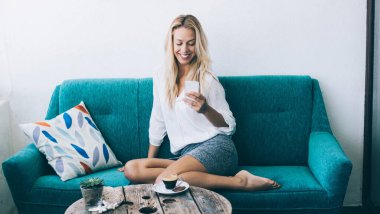 Gülümseyen milenyum kızı oturma odasındaki kanepede dinleniyor ve sosyal ağ kurarken cep telefonuyla e-posta yazıyor, neşeli kadın blogcu hafta sonu evde internet iletişiminin keyfini çıkarıyor.