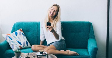 Neşeli hipster blogcusu sosyal ağlardan web takipçileriyle mesajlaşırken mutlu beyaz kadın sohbet etmek ve gülmek için cep telefonunu kullanarak rahat kanepede dinleniyor.