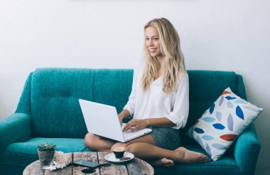 Uzaktan kumanda için kameraya gülümseyen neşeli yetenekli serbest yazar portresi, rahat koltukta dizüstü bilgisayarı olan beyaz kadın metin yazarı.
