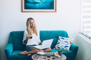 Uzaktan kumandalı beyaz iş hayatının tadını çıkaran mutlu Kafkasyalı, rahat koltukta modern bir dizüstü bilgisayarla oturuyor, milenyum kadınının, kahve molası sırasında kameraya gülümseyen portresi.