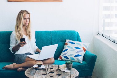 Uzaktan kumandalı ev dairesinde çalışan ve elinde akıllı telefon cihazıyla rahat bir koltukta oturan düşünceli sarışın bir kadın.