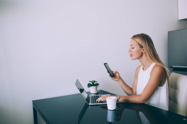 Çalışkan, sakin, şık elbiseli, akıllı telefonlu, ekrana odaklanan ve netbook 'la etkileşime giren bir iş kadını, çağdaş ofiste aromalı bir bardak sıcak içecekle masada oturuyor.