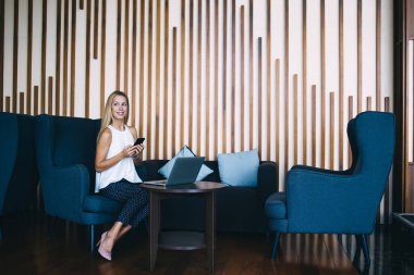 Tablet 'in yanındaki koltukta oturan ve cep telefonu kullanırken yüzünü çeviren tam boy sırıtkan kadın.