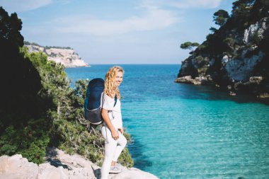 İspanyol adalarını keşfetmek için yapılan nefes kesici seyahat sırasında kameraya gülümseyen neşeli kadın turistin tam boy portresi sırt çantasıyla kayalık uçurumda poz veren mutlu Kafkasyalı gezgin.