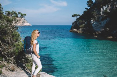 Menorca 'daki turkuaz su körfezinin nefes kesici manzaralı kayalık uçurumunu hayal eden kadın yürüyüşçü, turistik sırt çantalı kaygısız kadın solo gezi sırasında güzel doğa manzarasına hayran.