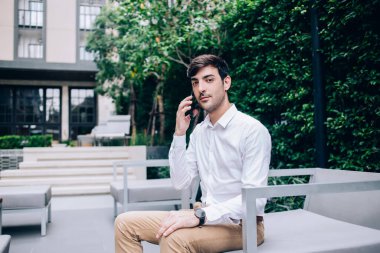 Şık giyinen düşünceli etnik erkek yönetici telefonda konuşuyor ve terasta koltukta oturup kameraya bakarken iş konularını tartışıyor.