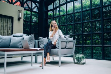 Şık giyinen memnun bayan yönetici cep telefonuyla konuşuyor. Masanın yanında dizüstü bilgisayarla oturuyor ve başka tarafa bakıyor.