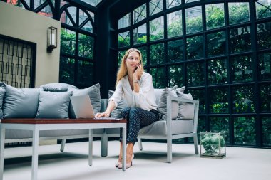 Şık giyinmiş neşeli bir bayan girişimci cep telefonuyla konuşuyor ve kafede çalışırken dizüstü bilgisayarda yazıyor ve başka yere bakıyor.