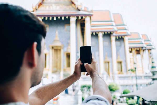 Back view of blurred man using smartphone to take photo of facade of ancient oriental palace while travelling in Bangkok Thailand