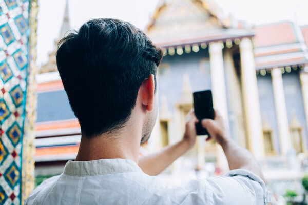 Back view of male tourist taking picture of historic landmark on mobile phone camera while sightseeing on city street during vacation in Bangkok