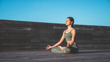 Nilüfer pozisyonunda, elleri dizlerinde, gözleri tahta kaldırımda kapalı, sakin, etnik kadın yoginin yan görünüşü.