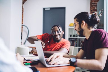 Gülümseyen Afrikalı Amerikalı bir adam, serbest çalışan meslektaşının yanında otururken ve internet hakkında bilgi ararken ve iş projesiyle ilgili fikirleri tartışırken dizüstü bilgisayar ekranını işaret ediyor.