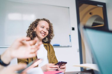 Çağdaş ofisteki iş arkadaşlarıyla iş görüşmesi sırasında masada otururken gülümseyen ve akıllı telefonla konuşan neşeli kıvırcık kadın.